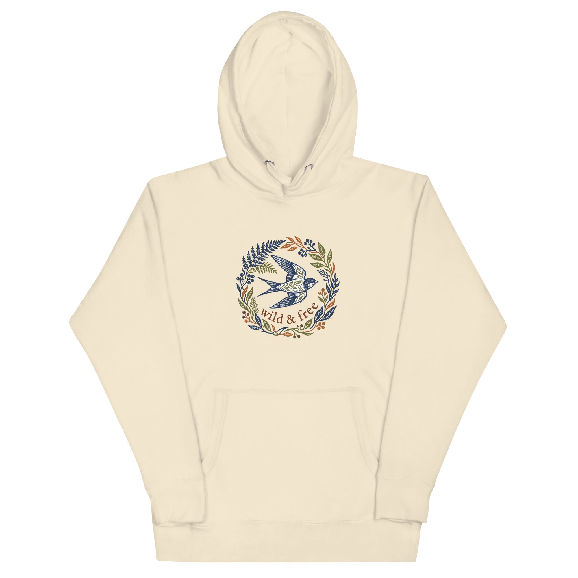A light bone beige pullover hoodie laid flat against a white background. The front features a central graphic design of a stylized flying swallow surrounded by a wreath of ferns, leaves, and berries in navy, olive, and rust colors. Below the bird, the text "wild & free" is written in a brown serif font.