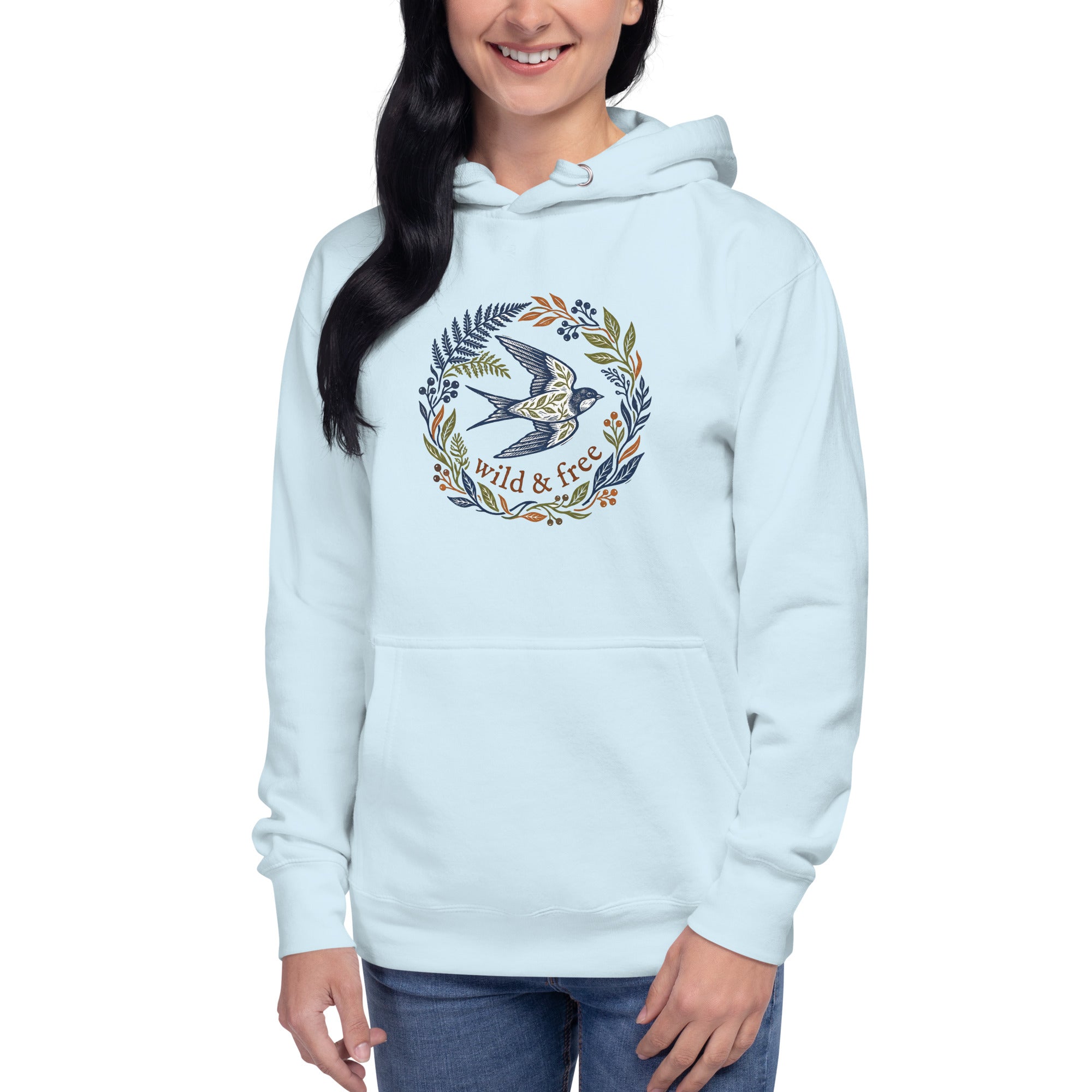 A smiling woman with long dark hair modeling the sky-blue hoodie. She is standing facing forward, wearing blue jeans. The "wild & free" bird and floral graphic is clearly visible on the center of the chest.