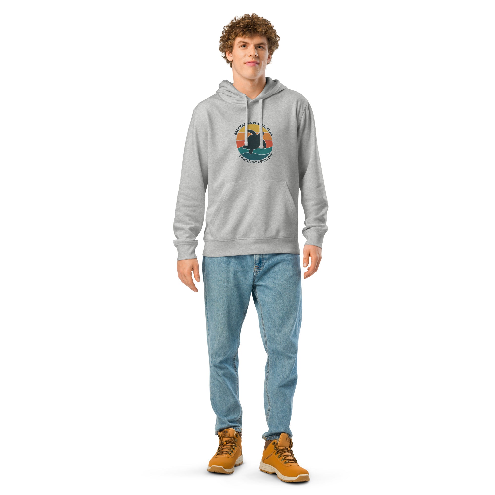 A young man with curly light brown hair stands against a white background wearing a light heather grey hoodie, light-wash blue jeans, and tan work boots. The hoodie features a circular graphic on the chest with a sea turtle silhouette and the text "Keep the sea plastic free - Earth day every day."