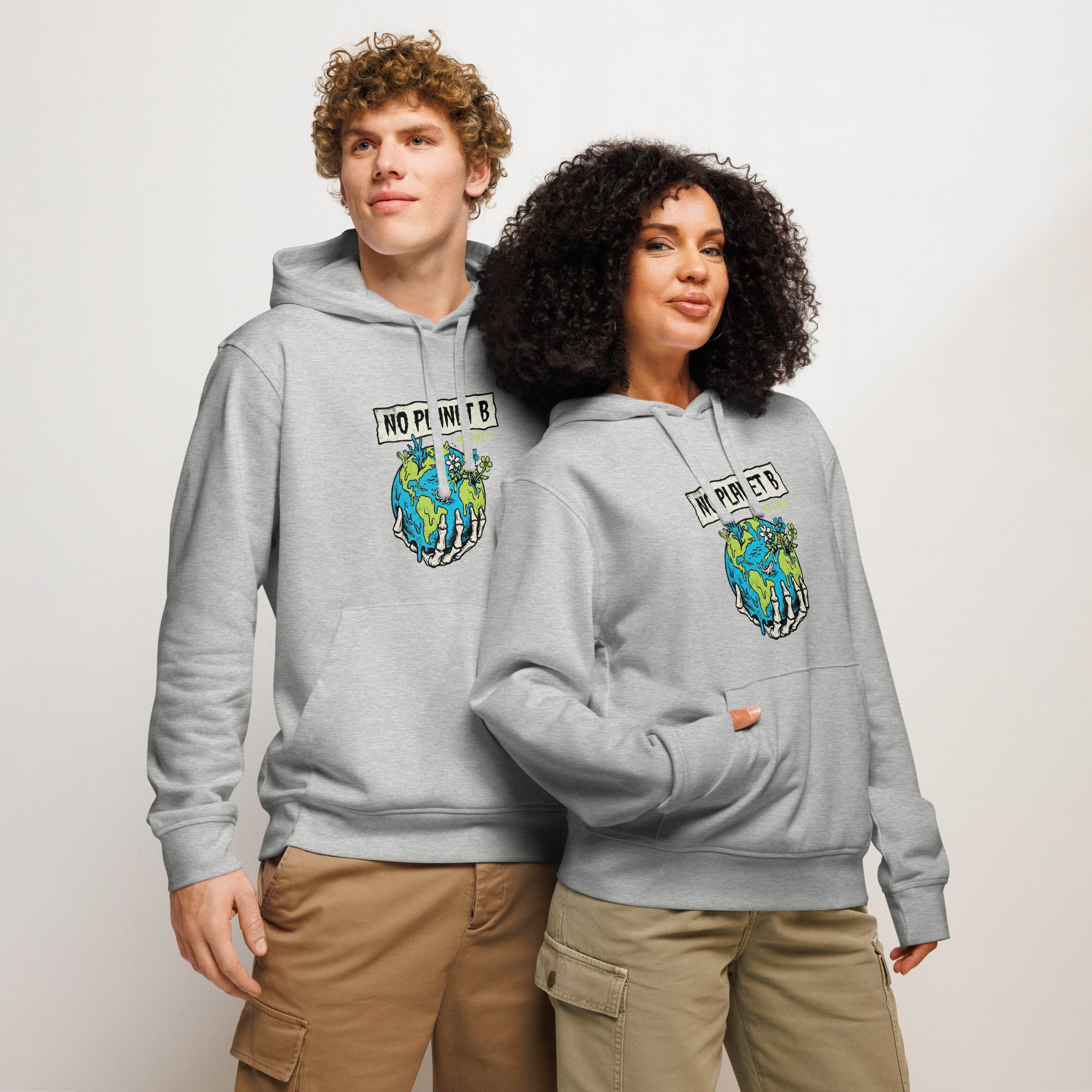 Two people stand side-by-side against a light background wearing matching light heather grey "No Planet B" hoodies. One person has light curly hair and wears tan cargo pants, while the other has dark voluminous curly hair and wears olive green cargo pants. Both hoodies display the skeletal hand and Earth graphic on the chest.