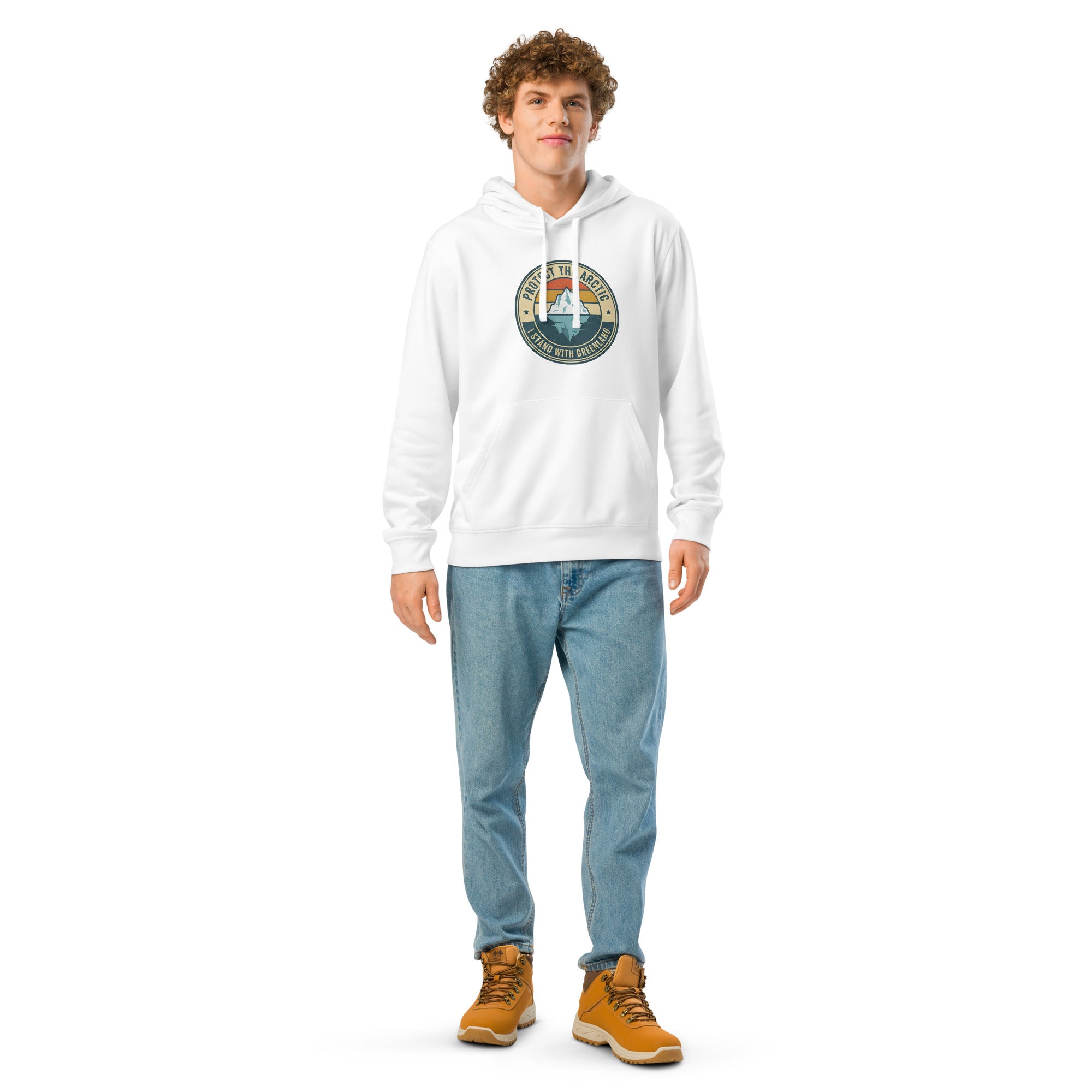 A man wearing a white hoodie featuring a circular vintage-style graphic on the chest with an iceberg, sunset-colored stripes, and the text "PROTECT THE ARCTIC" and "I STAND WITH GREENLAND," paired with light-wash jeans and brown boots.