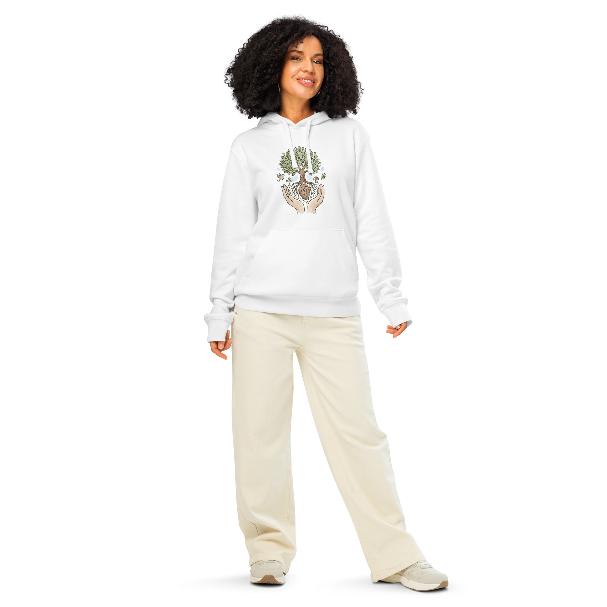 A smiling person with dark curly hair standing in a full-length pose. They are wearing the white "Tree of Life" hoodie paired with off-white wide-leg trousers and neutral sneakers.