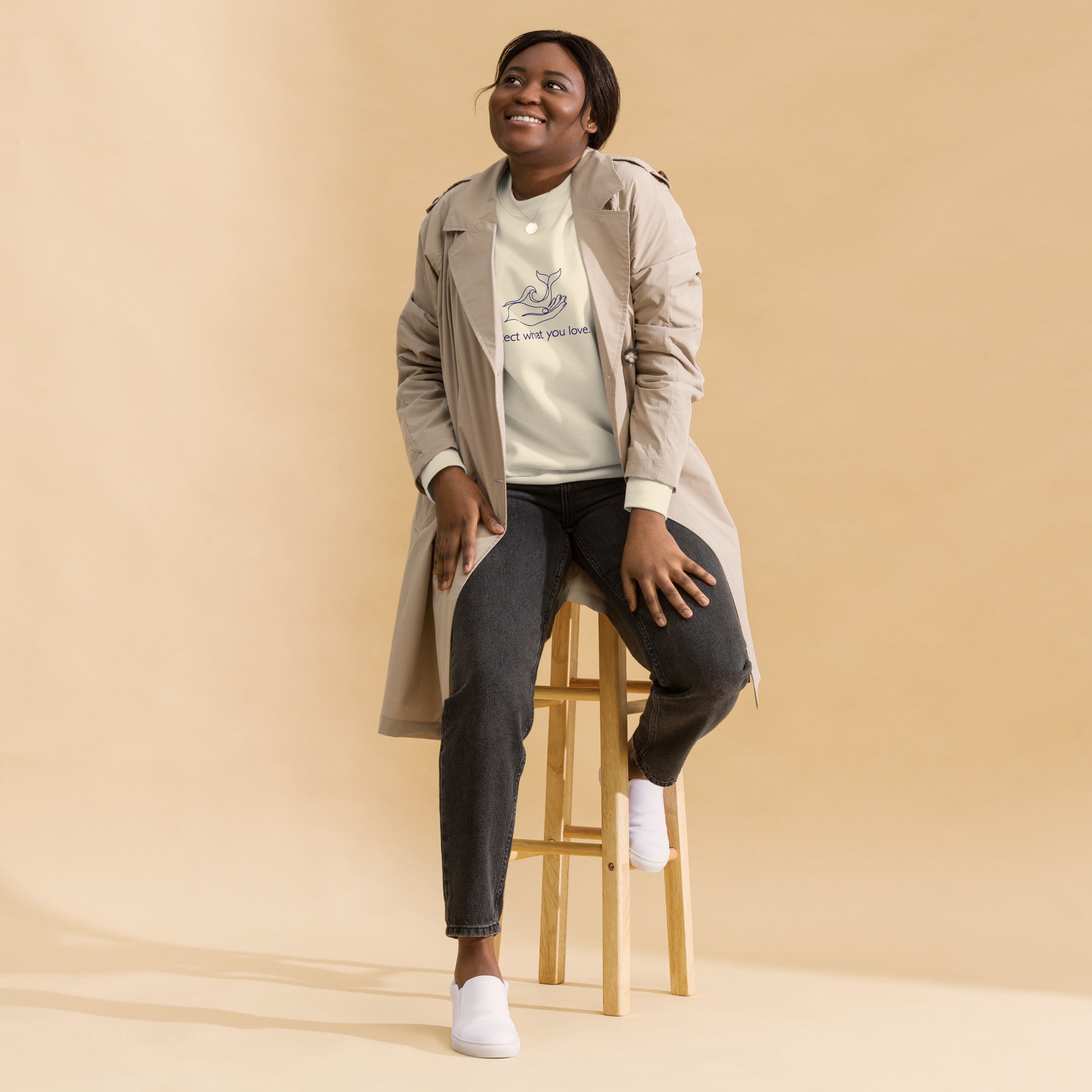 A person sitting on a wooden stool against a tan background, wearing the bone-colored "Protect What You Love" sweatshirt. They are layered in a tan trench coat, dark grey jeans, and white slip-on shoes, looking upward with a smile.