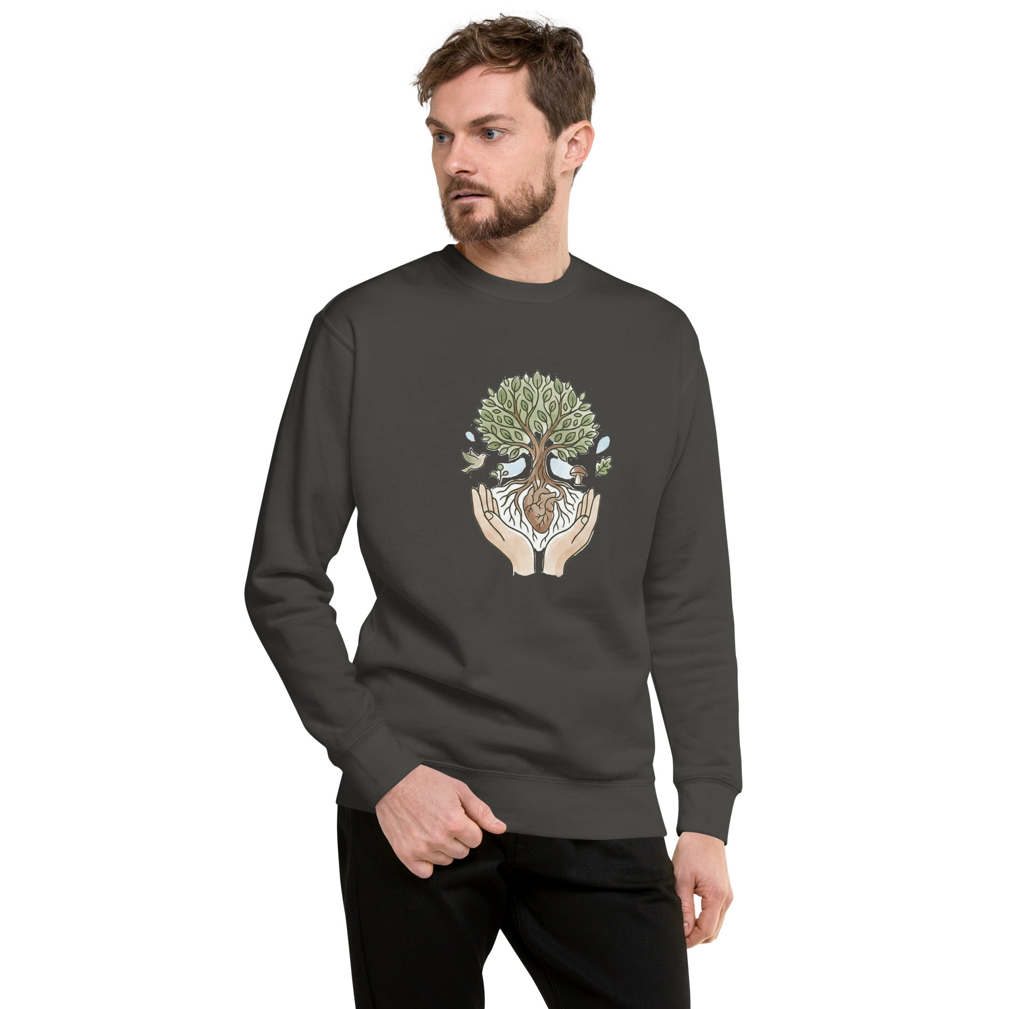 A man with a short beard and brown hair is shown from the waist up, looking away from the camera. He is wearing a dark charcoal grey crewneck sweatshirt featuring a central graphic of a leafy tree cradled by two hands. The tree's roots are intertwined around a small brown heart, and a bird and a mushroom are illustrated near the base. He has his right hand in his pocket and is wearing black trousers.