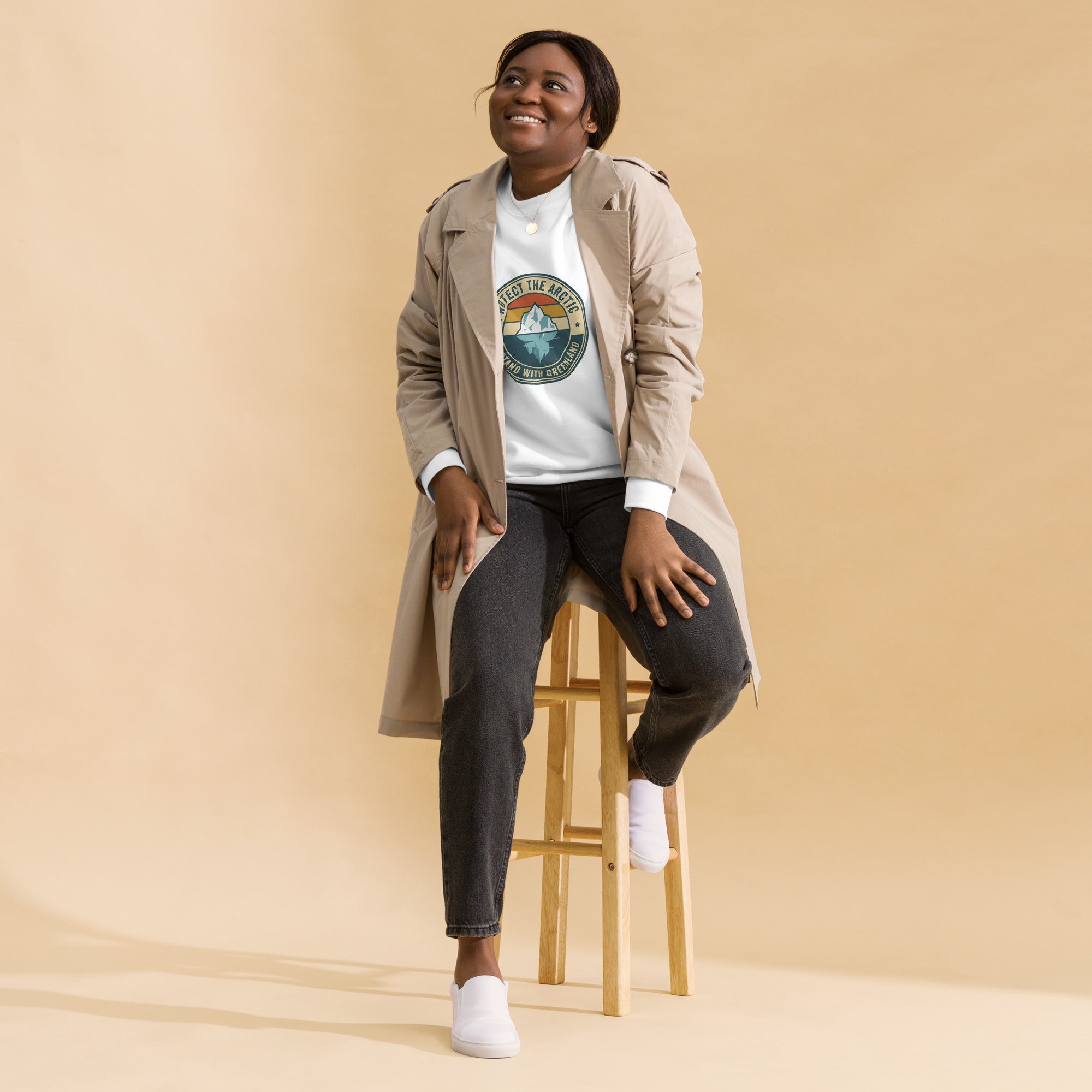 A person sitting on a wooden stool wearing a white sweatshirt that features a vintage-style circular graphic of an iceberg with the text "PROTECT THE ARCTIC" and "I STAND WITH GREENLAND," layered under a tan trench coat.