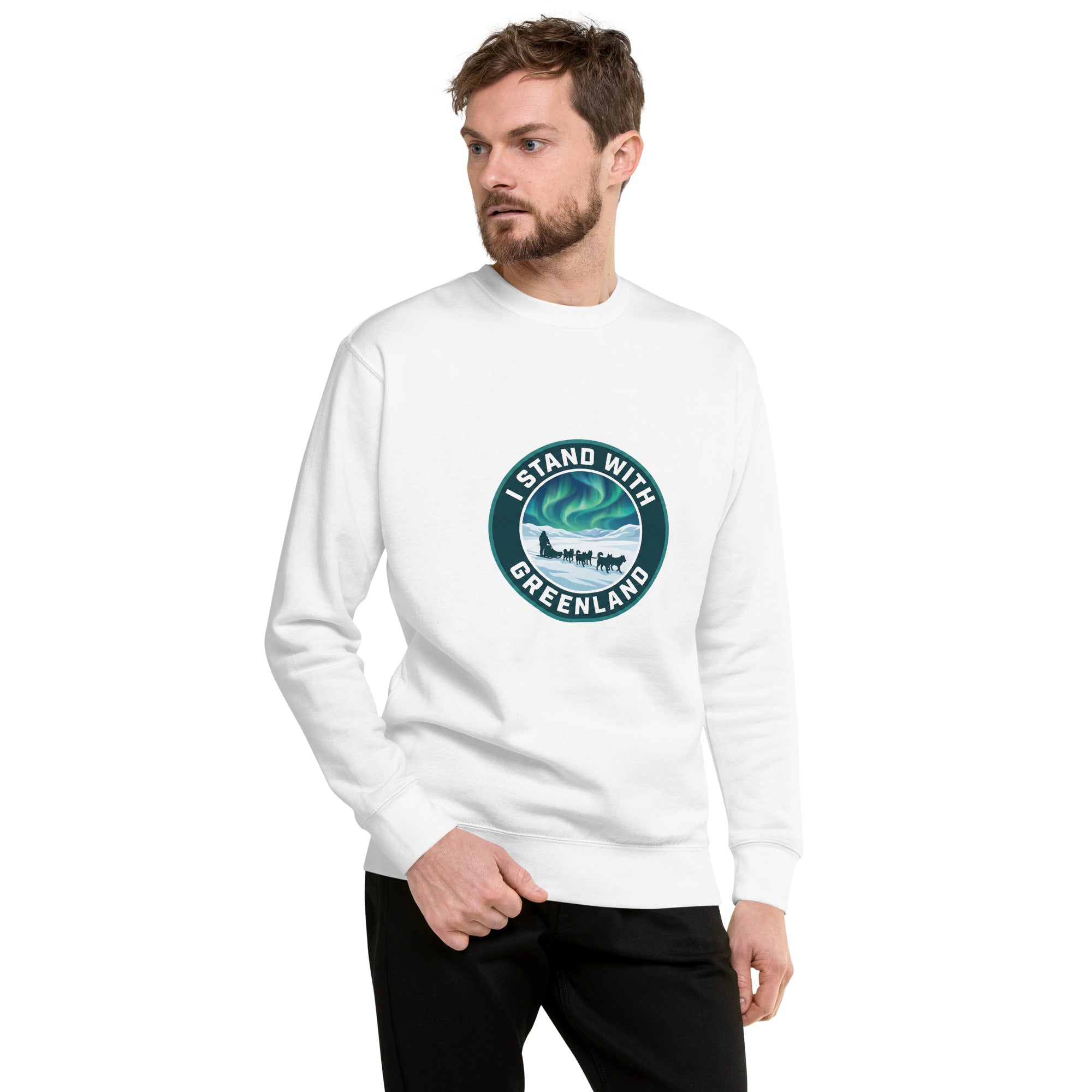A man with a beard is looking to his right while wearing a white long-sleeved sweatshirt. The sweatshirt features a circular teal graphic on the chest depicting a dog sled team traveling under the green Northern Lights, surrounded by the text "I STAND WITH GREENLAND". He is standing with one hand in the pocket of his black trousers against a plain white background.